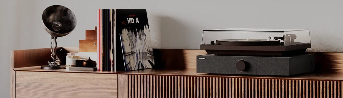 Black SpinBase 2 and turntable on a sideboard beside records and a lamp.