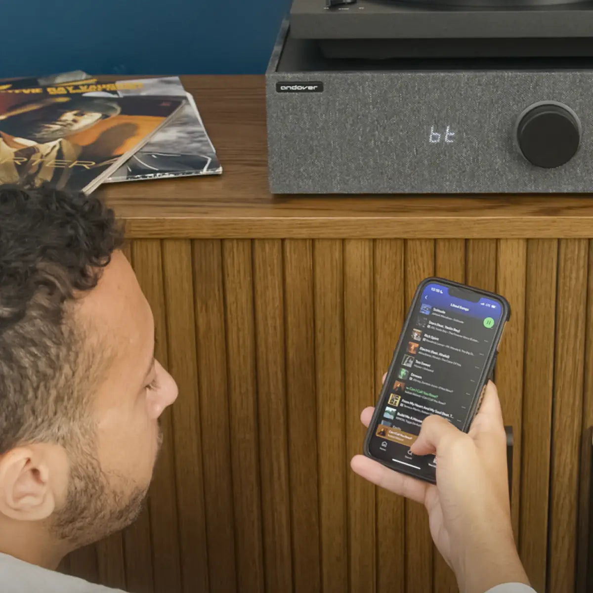 Listener selecting music on a smartphone beside a SpinBase MAX 2 displaying Bluetooth mode.